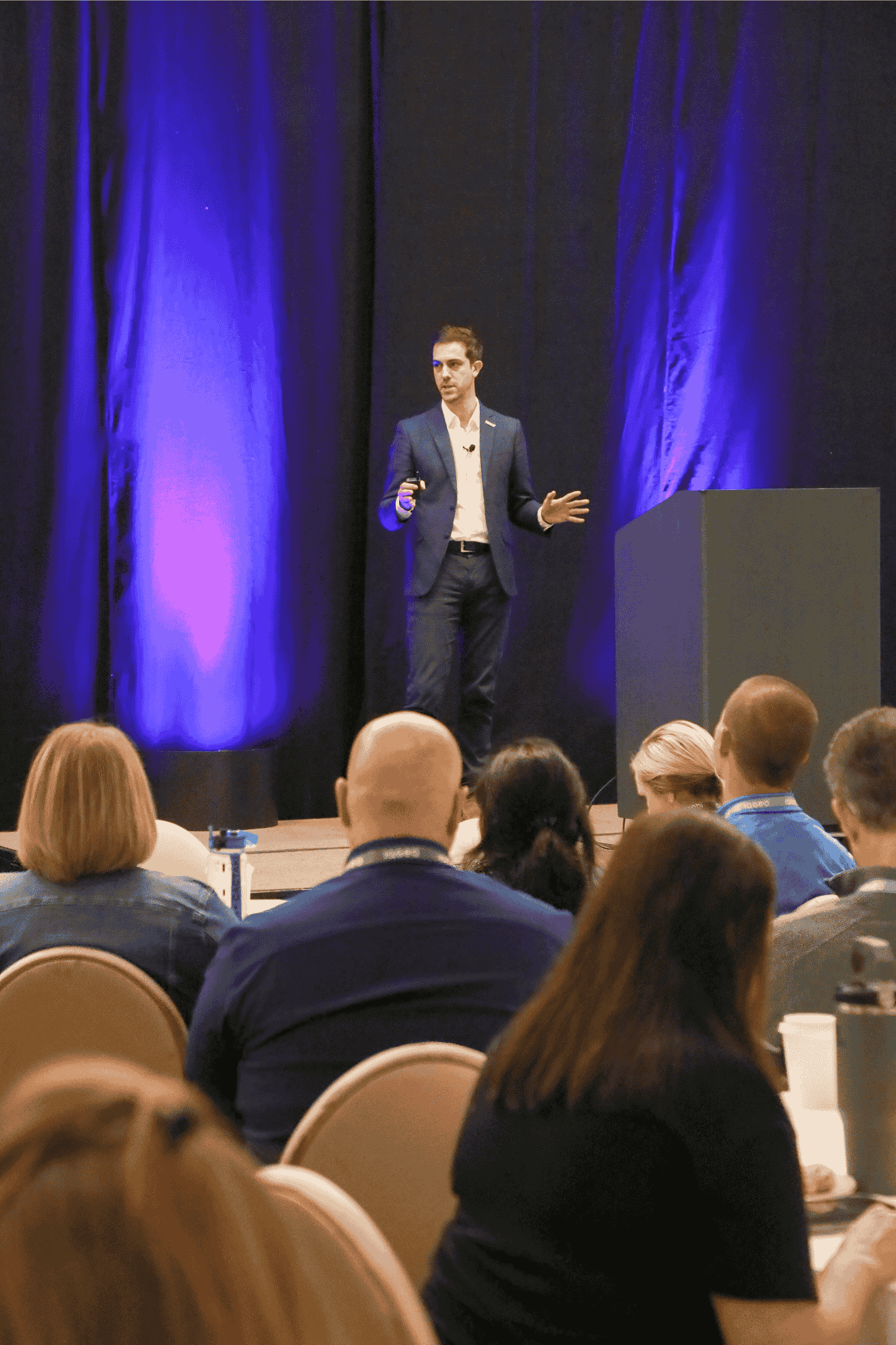 Aloïs Brunel from IQGeo presenting on stage at the IQGeo Americas Meetup 2025 in Dallas, addressing a full audience under purple stage lighting