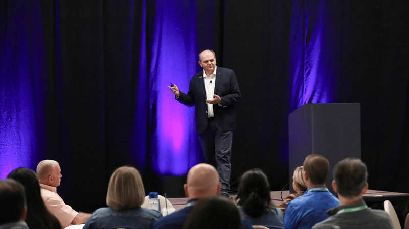 IQGeo CEO, Richard Petti, presenting on stage at the IQGeo Americas Meetup 2025 in Dallas, addressing a full audience under purple stage lighting