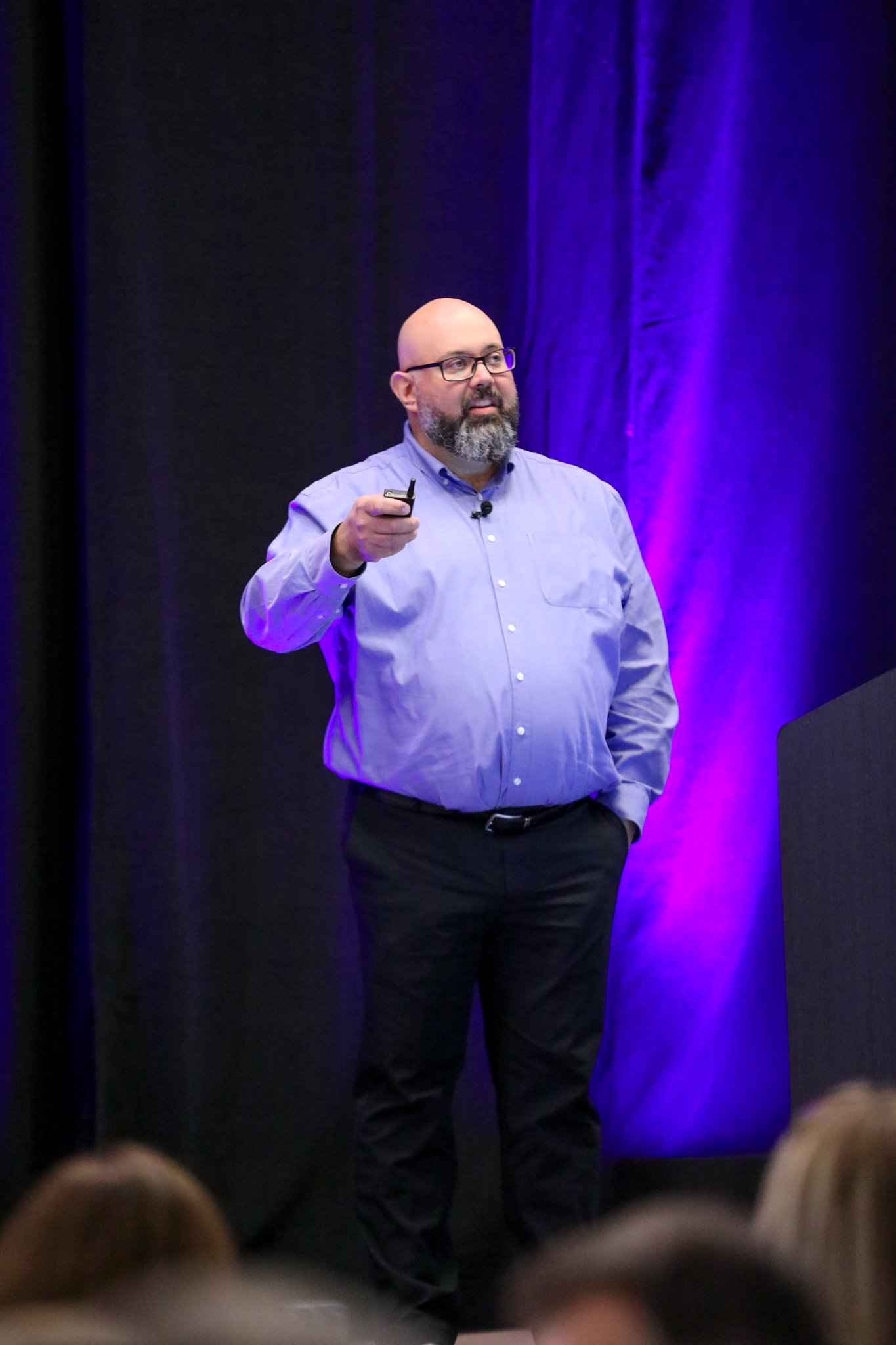 Jason Lemon from SaskTel presenting on stage at the IQGeo Americas Meetup 2025 in Dallas, addressing a full audience under purple stage lighting