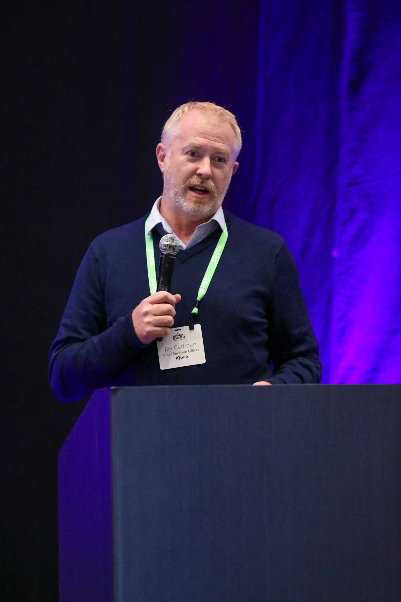 Jay Cadman from IQGeo presenting on stage at the IQGeo Americas Meetup 2025 in Dallas, addressing a full audience under purple stage lighting