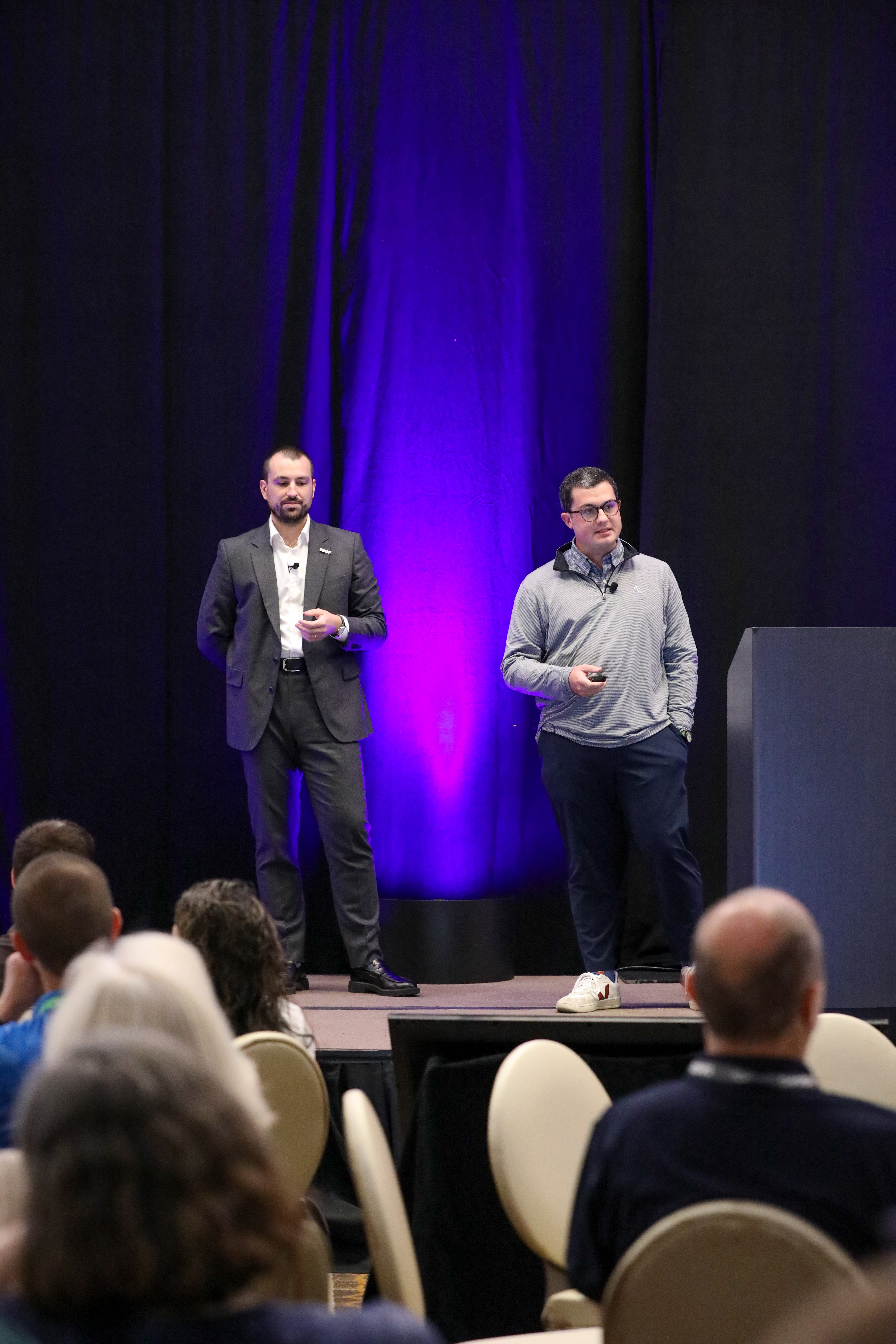 Thomas Thuillier and Matt Roberts from IQGeo presenting on stage at the IQGeo Americas Meetup 2025 in Dallas, addressing a full audience under purple stage lighting