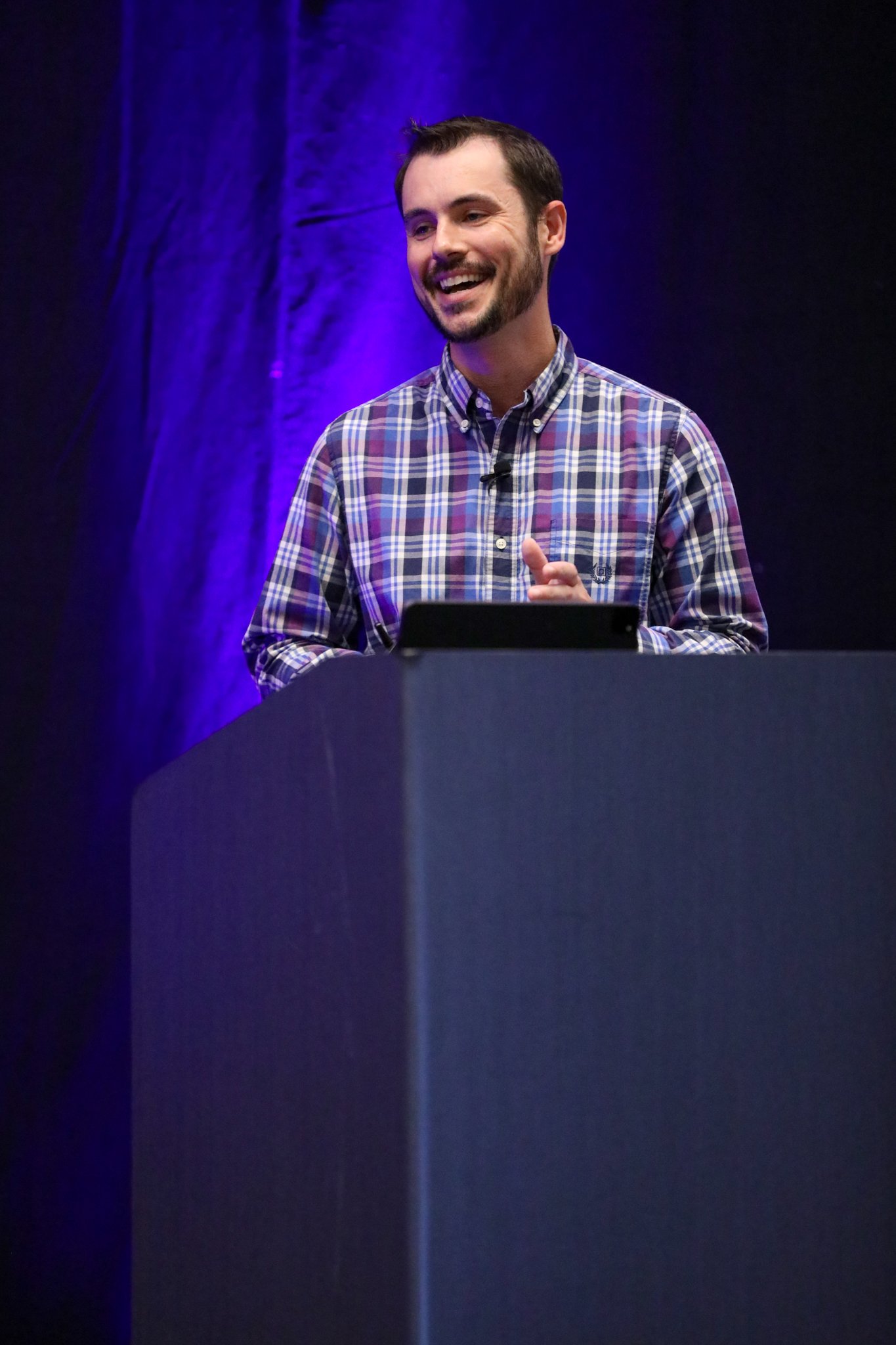 Ryan Cox from PPL presenting on stage at the IQGeo Americas Meetup 2025 in Dallas, addressing a full audience under purple stage lighting