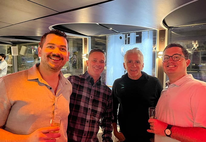 Four people stand together indoors at a social event, each holding a drink, with warm lighting and modern décor in the background.