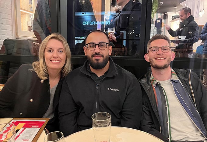 Three people sit together at a café table with drinks, posing for a photo indoors