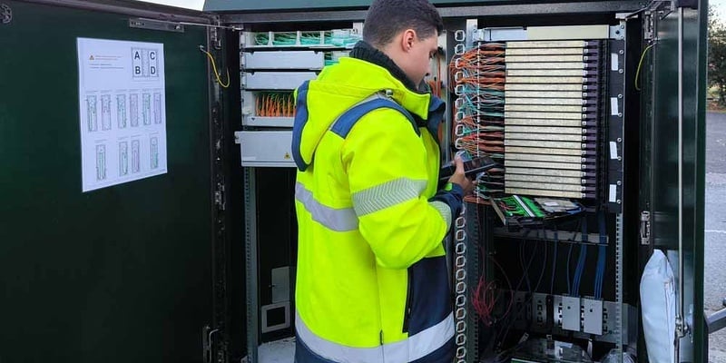 Telecom field technician in hi-vis jacket connecting cables inside an open telecom cabinet.
