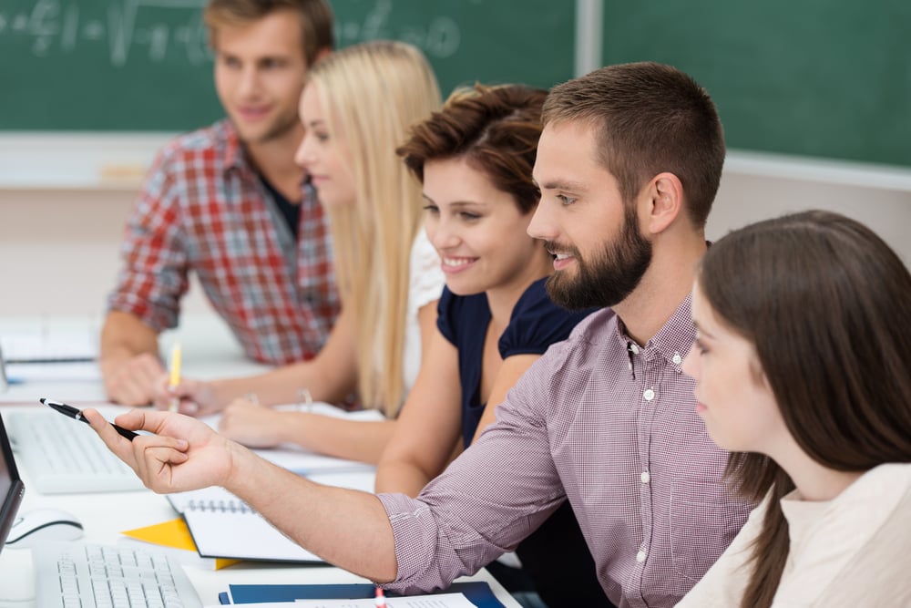 Diverse group of attractive young university students studying together sitting at a table sharing a computer monitor