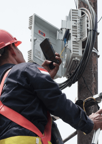 A utility worker checks equipment on a pole while a laptop displays a project dashboard with inspection photos and metrics.