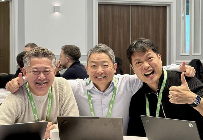 Three people sit together at a table with laptops during a meeting or workshop, with one giving a thumbs‑up.