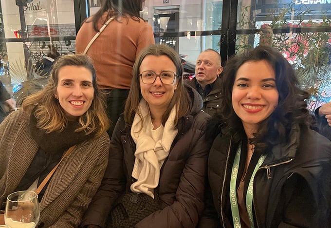 Three people sit together at a café table, bundled in winter clothing, with warm indoor lighting and a city street visible through the window behind them.