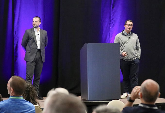 Two presenters stand on stage in front of a purple-lit backdrop, one speaking while the other listens