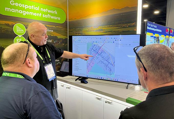 A presenter points to a large screen showing a geospatial map while two people watch at a trade show booth.