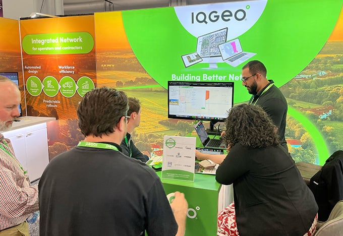  People gather at an IQGeo trade show booth where a presenter demonstrates software on a monitor.
