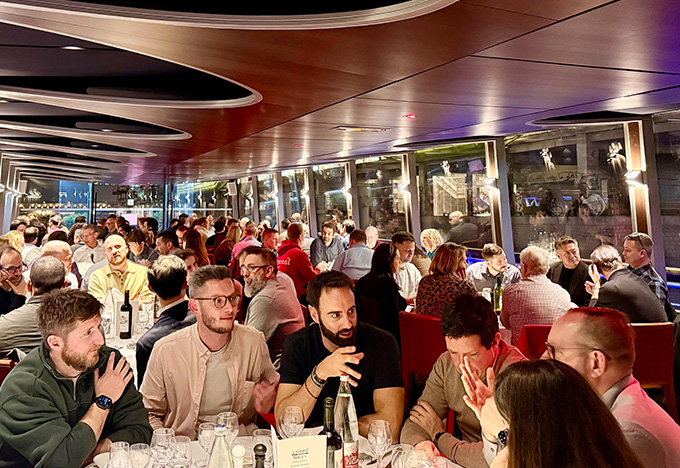 A large group of people sit closely together at tables during a lively dinner event on a boat